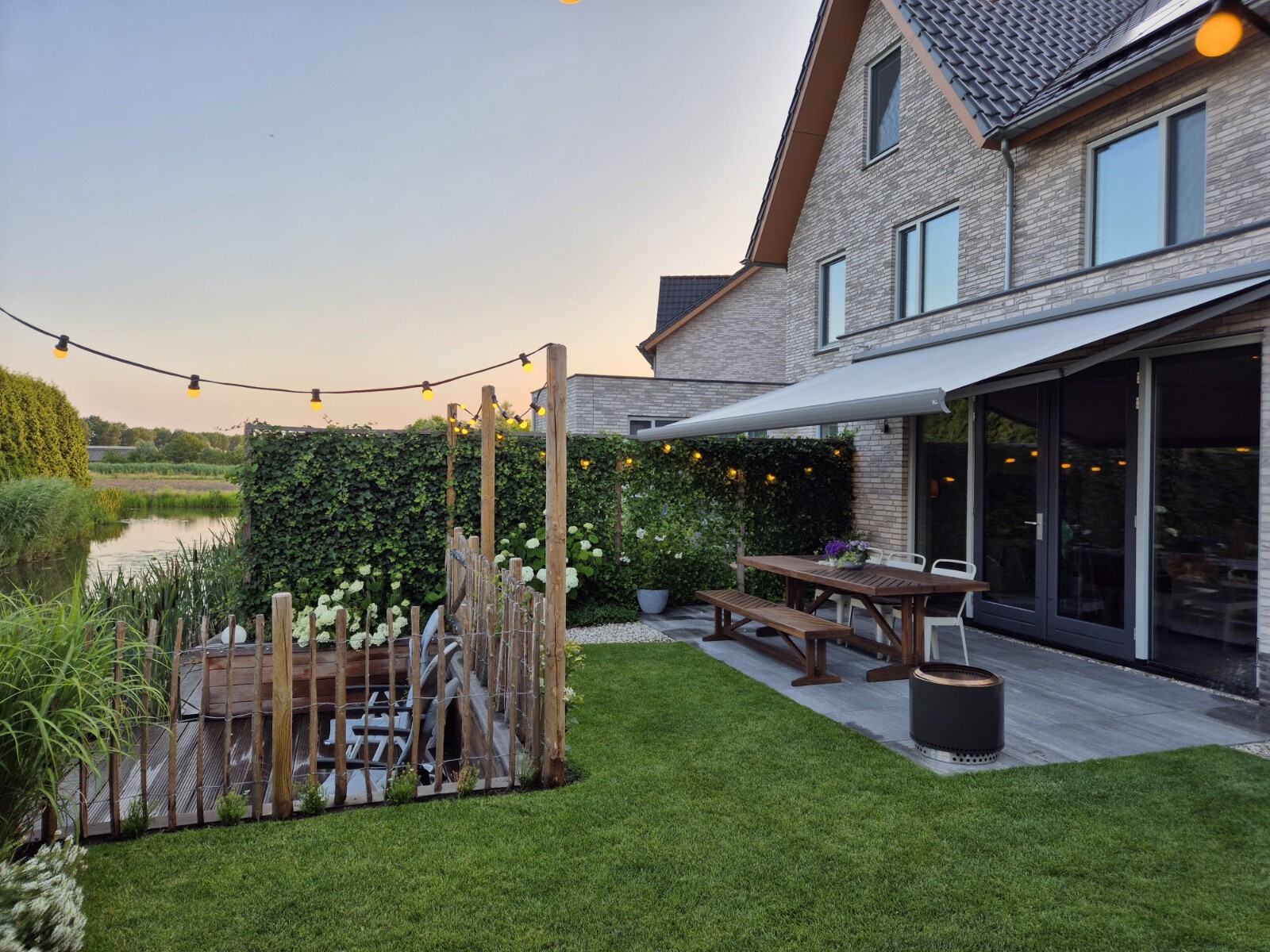 Knikarmscherm als elektrische zonwering voor buiten in een tuin met gazon aan een watertje.